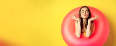 Beautiful coquettish woman inside pink swimming ring, posing on summer vacation, beach holiday concept, standing against yellow background.