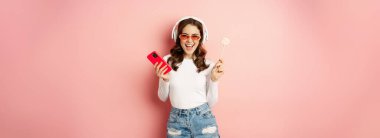 Dancing woman in sunglasses, listening music in headphones, holding lolipop and smartphone, laughing and smiling happy, standing over pink background.