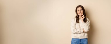 Smiling young woman thinking, planning smth, making choice and looking up with thoughtful face, beige background.