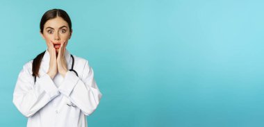 Portrait of shocked woman doctor, female hospital intern in white coat, looking concerned and confused at camera, disbelief face, standing over torquoise background.