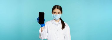 Angry and confused woman doctor in face mask, gloves, showing smartphone app, mobile screen, frowning upset, standing over blue background.