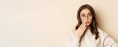 Close up portrait of girl in glasses looking surprised, say oops, standing coquettish over beige background. Copy space