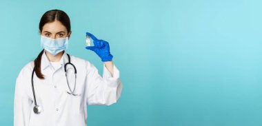 Aşı kampanyası. Yüzünde tıbbi maske ve eldiven olan gülümseyen kadın doktor Covid, Omicron delta varyantı aşısı gösteriyor. Torkuaz arka planda duruyor..