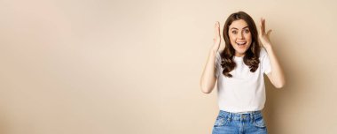 Enthusiastic adn surprised woman winning, dancing and celebrating, standing in tshirt over beige background.