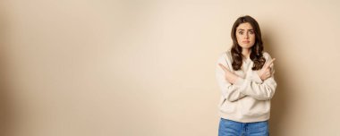 Complicated and indecisive woman pointing sideways, cant make choice, struggle with decision, choosing between two options, standing over beige background.