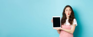 Şaşırmış çekici bir kadın dijital tablet ekranı gösteriyor ve kameraya hayret içinde bakıyor, tanıtım teklifini kontrol ediyor, mavi arka planda eğleniyor..