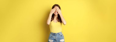 Portrait of brunette woman cover eyes with hands, blindfolded, smiling with anticipation, expecting smth, waiting for surprise, standing over yellow background.