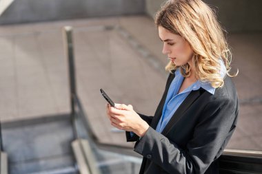 Yürüyen merdivendeki genç iş kadınının portresi, akıllı telefon ve gülümseme kullanarak, cep telefonunu dinleyerek, takım elbise giyerek..
