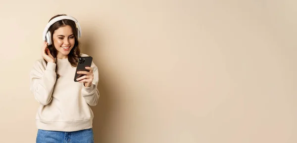 Beautiful young woman looking at video on smartphone, listening music in headphones, standing in casual clothes over beige background.