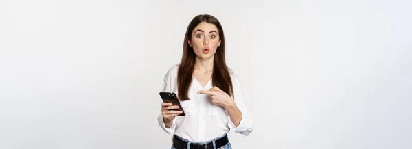 スマホで笑顔を見せながらアプリに興味を持ちながら携帯電話を指している若い女性が白い背景の上に立っている — ストック写真