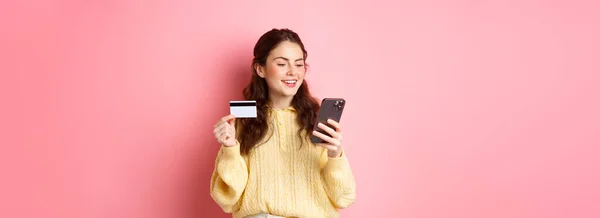 技术和网上购物 年轻漂亮的女士在线支付信用卡 看着智能手机和微笑 站在粉红的背景之上 — 图库照片