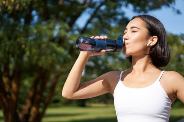 Gülümseyen Asyalı fitness kızı su içer, parkta egzersiz yapar, koşu antrenmanı sırasında susuz kalmaz..