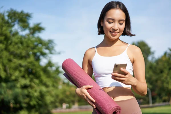 年轻的亚洲女人在运动时检查手机 拿着橡皮垫在公园里散步 去健身房 拿着智能手机的肖像 — 图库照片