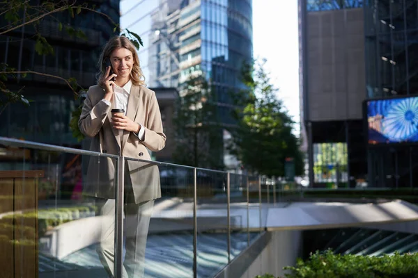 stock image Stylish office woman in beige suit, talking on mobile phone while standing outside on street with morning coffee.