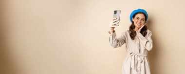 Beautiful girl taking selfie on smartphone in coat and cute hat, smiling for photo and looking happy, standing over beige background.