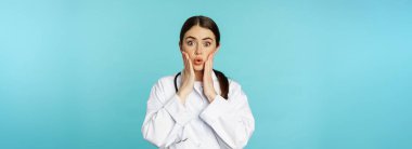 Portrait of shocked female doctor, worried healtcare worker in white robe, looking concerned at patient, standing over blue background.