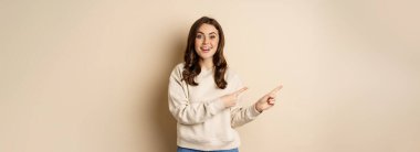 Cheerful attractive female model pointing fingers left, showing store advertisement, banner or logo, standing in sweater over beige background.