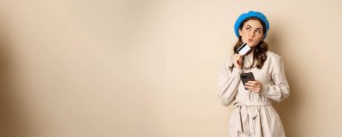 Portrait of trendy woman in coat holding smartphone, showing credit card, shopping online, order something on mobile phone, standing over beige background.
