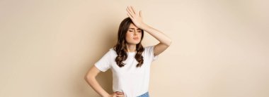 Disappointed woman reacting on failure, slap forehead, facepalm with sad face, beige background.