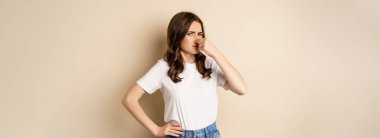 Woman shut nose from disgusting smell, standing over beige background.
