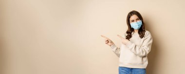 Health and covid-19 concept. Confused young woman in medical face mask, pointing left and staring with frustrated face, beige background.