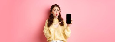 Teknoloji ve online alışveriş. Heyecanlı kız meraklı görünüyor, boş cep telefonu ekranını işaret ediyor, akıllı telefondan uygulamayı gösteriyor, pembe arka planda duruyor..