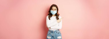 Beautiful stylish woman in medical face mask from covid, pointing finger at upper left corner, showing advertisement, standing over pink background.