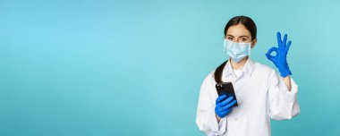 Online doctor and clinic. Young woman in medical face mask, using smartphone for client remote online appointment, showing okay sign, standing over blue background.