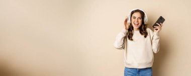 Happy caucasian woman listening music in headphones on smartphone app, dancing and having fun, standing over beige background.
