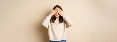 Excited happy girl smiling, close eyse and wait for surprise gift, standing blinsided over beige background.