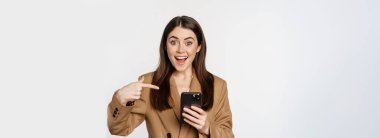 Enthusiastic saleswoman, business woman pointing finger at mobile phone and smiling, showing on cellphone, standing against white background.