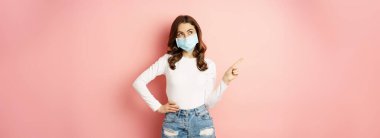Beautiful stylish woman in medical face mask from covid, pointing finger at upper left corner, showing advertisement, standing over pink background.