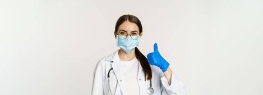 Close up portrait of woman doctor, physician in face mask from coronavirus, showing thumbs up and smiling, recommending clinic or medical website, white background.