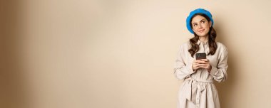 Dreamy smiling woman in stylish trenchcoat, looking up fantasizing while shopping on mobile phone app, using smartphone and thinking, standing over beige background.
