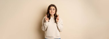 Hopeful worried girl cross fingers, making wish, standing over beige background.