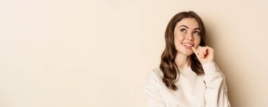 Cute modern woman thinking, daydreaming and smiling, looking up thoughtful, standing over beige background.