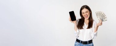 Enthusiastic young woman winning money, showing smartphone app interface and cash, microcredit, prize concept, standing over white background.