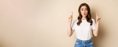 Excited girl looking interested, pointing fingers up, showing advertisement, standing in tshirt over beige background.