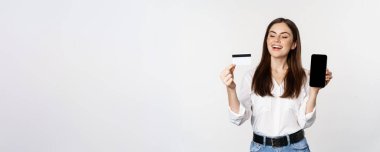 Happy woman showing credit card and smartphone screen, concept of online shopping, buying in app, standing over white background.
