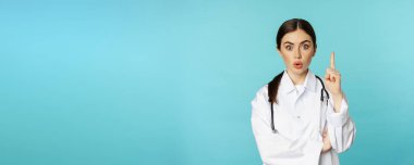 Young doctor, woman physician in white coat raising finger, pointing up, suggesting smth, has solution, revelation, standing over torquoise background.