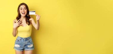 Portrait of brunette girl showing her credit card and smiling, wearing summer clothes, concept of bank, money and shopping, standing over yellow background.