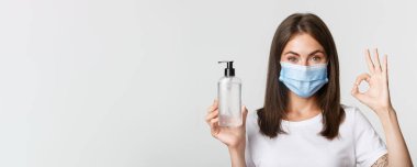 Covid-19, sağlık ve sosyal uzaklık kavramı. Tıbbi maskeli, el dezenfektanı ve iyi bir jest gösteren gülümseyen esmer kızın yakın çekimi..