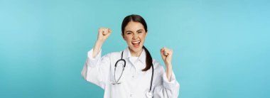 Enthusiastic doctor woman, rejoicing, winning and celebrating, achieve goal, standing in white coat against blue background. Copy space