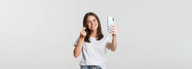 Mutlu, güzel, genç bir kadın kalp hareketi yapıyor, akıllı telefondan selfie çekiyor ve gülümsüyor..