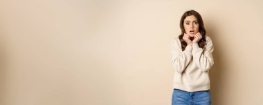 Scared insecure woman trembling from fear, looking at camera shocked and frightened, standing over beige background.