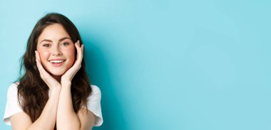 Güzel bir kadının parıldayan tenine dokunduğu, makyaj yaptığı, dişleriyle gülümsediği, mavi arka planda dikildiği yakın plan..