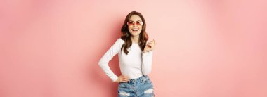 Portrait of stylish glam girl in sunglasses, laughing and smiling, standing over holiday pink background. Copy space