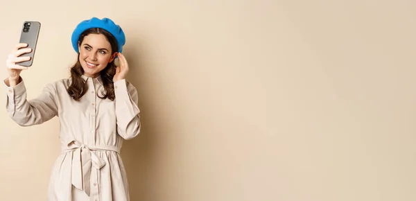 stock image Beautiful girl taking selfie on smartphone in coat and cute hat, smiling for photo and looking happy, standing over beige background.