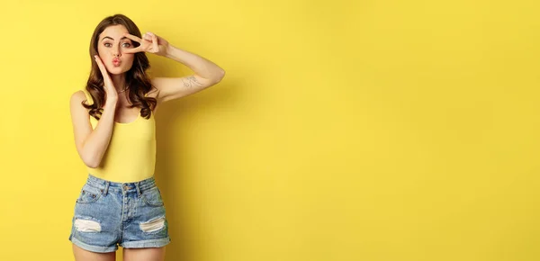 stock image Beautiful glamour girl in summer outfit, showing peace sign, kissing face, standing against yellow background.