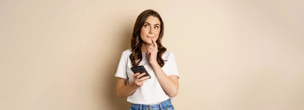 Girl Thinks Holds Mobile Phone Looks Thoughtful Stands Beige Background — Zdjęcie stockowe